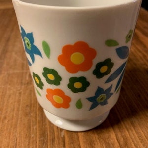 May include: White ceramic mug with a floral design. The flowers are orange, yellow, blue, and green.
