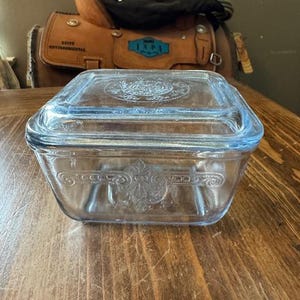 Peut inclure: Un plat à casserole rectangulaire en verre bleu clair vintage avec un couvercle. Le plat présente des motifs floraux et de volutes en relief. Le couvercle et le plat ont une forme légèrement arrondie. Le plat est posé sur une surface en bois.