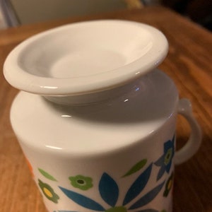 May include: White ceramic mug with a floral design featuring blue, green, orange, and yellow flowers. The mug has a wide rim and a handle.