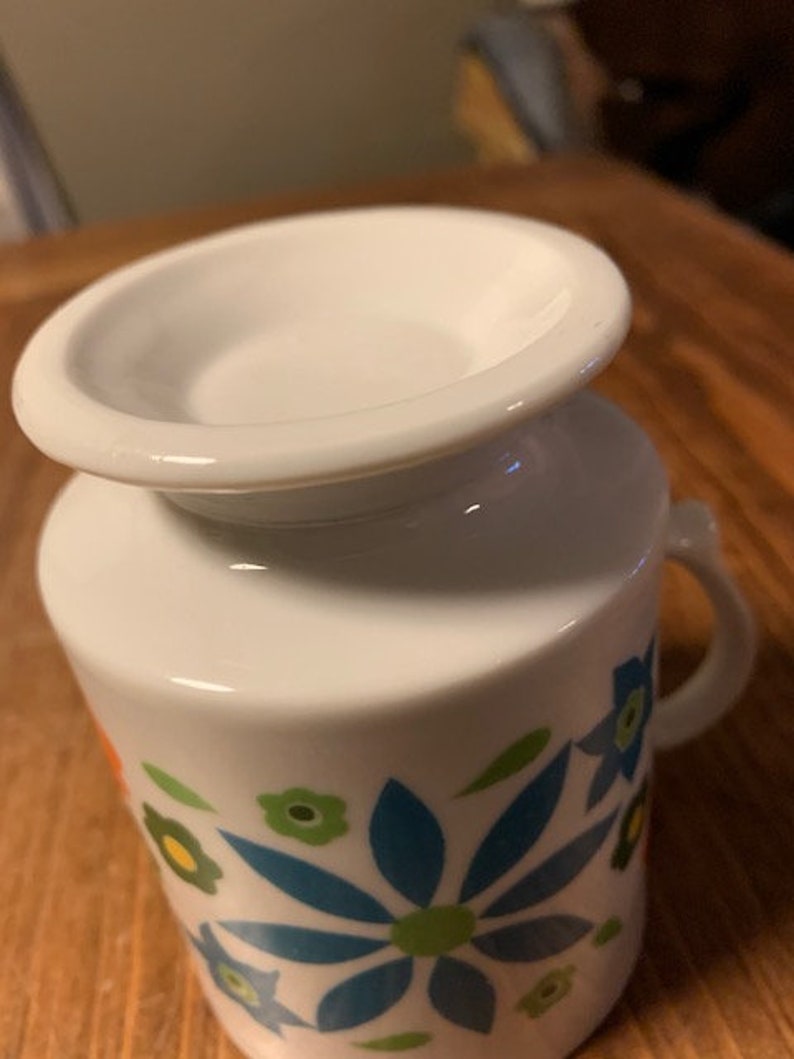 May include: White ceramic mug with a floral design featuring blue, green, orange, and yellow flowers. The mug has a wide rim and a handle.