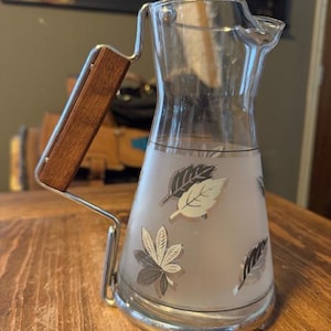 Op de afbeelding: Een heldere glazen kan met een zilveren voet en handvat. De kan heeft een houten handvat en is versierd met bladeren in grijstinten en wit. De kan staat op een houten tafel.