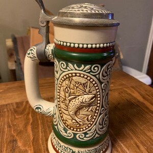 May include: A white ceramic beer stein with a green and red design. The stein features a detailed image of a fish and the text "The Strike. Rainbow Trout."