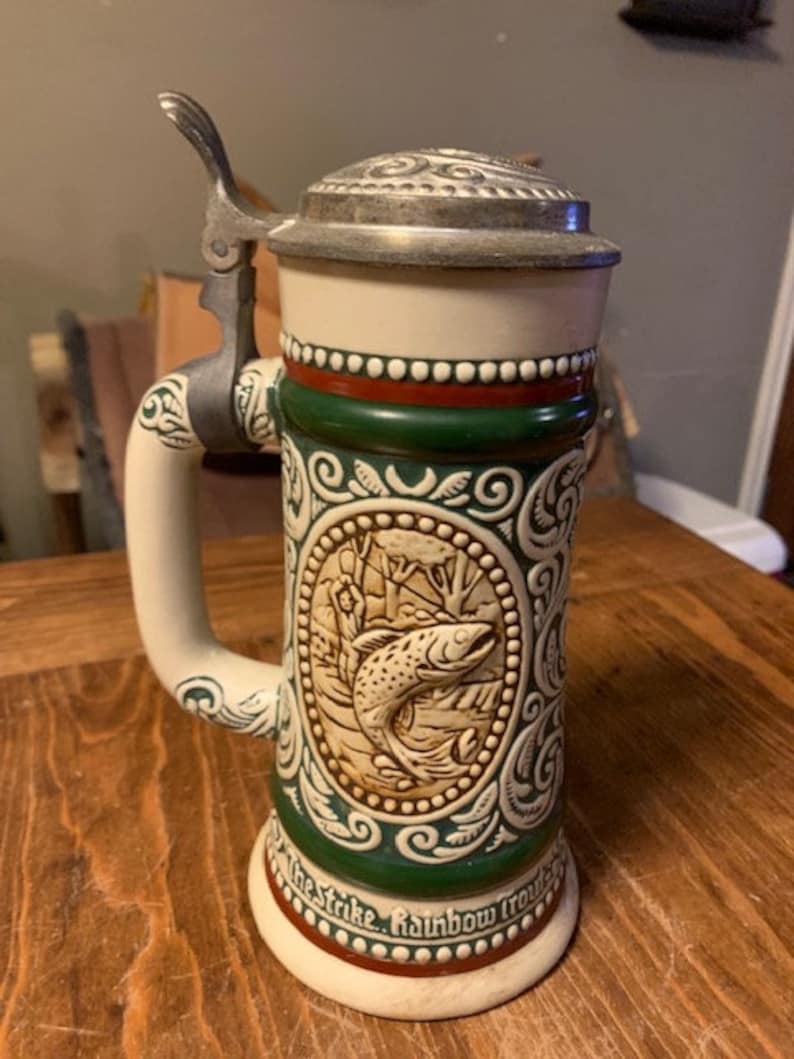 May include: A white ceramic beer stein with a green and red design. The stein features a detailed image of a fish and the text "The Strike. Rainbow Trout."