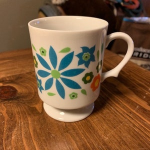May include: White ceramic mug with a footed base. The mug has a floral design with blue, green, yellow, and orange flowers.