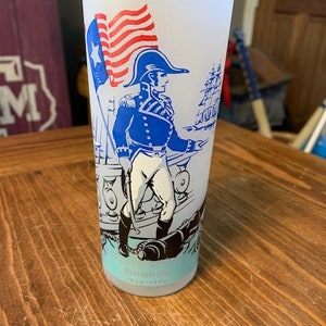 Könnte beinhalten: Ein weißes, gefrostetes Glas mit einer blauen und roten Illustration eines Mannes in Militäruniform, der eine amerikanische Flagge hält und vor einem Schiff steht. Der Text "U.S. Navy 1775-1975" ist auf der Unterseite des Glases aufgedruckt.
