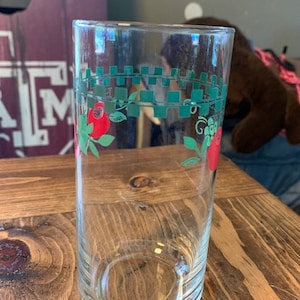 May include: A clear glass drinking glass with a green and red design. The design features a vine with red berries and green leaves. The vine is wrapped around the glass and is bordered by a green checkered pattern.