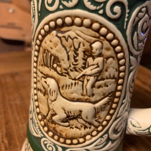 May include: A green and white ceramic beer stein with a raised design of a man and a dog hunting. The design is in brown and white and is surrounded by a green and white border.