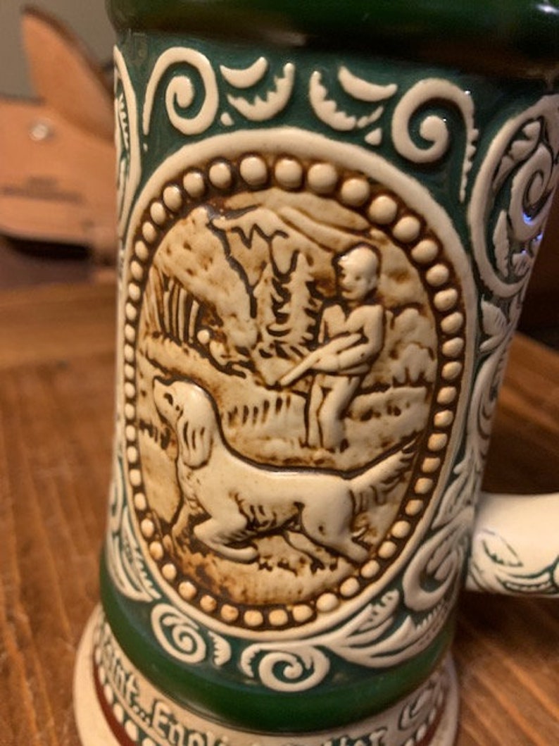May include: A green and white ceramic beer stein with a raised design of a man and a dog hunting. The design is in brown and white and is surrounded by a green and white border.