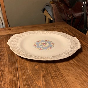 May include: A white ceramic serving platter with a gold rim and a floral design in the center. The platter has two handles on the sides.