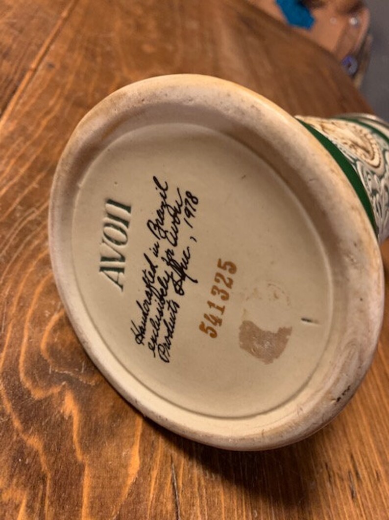May include: A white ceramic mug with a green and gold design. The bottom of the mug has the text "AVOII Handcrafted in Brazil Exclusively for C.W.P. Products Office, Inc. 1978 5411325"