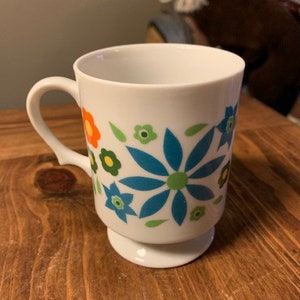 May include: White ceramic mug with a blue, green, orange, and yellow floral design. The mug has a handle and a pedestal base.