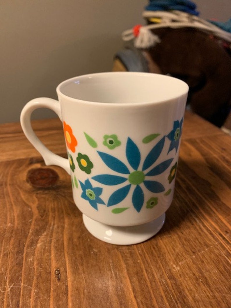 May include: White ceramic mug with a blue, green, orange, and yellow floral design. The mug has a handle and a pedestal base.