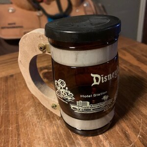 以下が含まれることがあります： 白いストライプと黒い蓋が付いた茶色のガラス製のマグカップ。マグカップには木製のハンドルと、ディズニーランドのホテルステーションの白いグラフィックが描かれています。マグカップには「ディズニーランド」の文字が印刷されています。