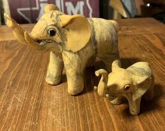 May include: Two decorative elephant figurines made of a light brown and white marbled material. The larger elephant is standing with its trunk raised, while the smaller elephant is standing with its trunk down. Both elephants have black eyes.