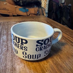 May include: A cream-colored soup bowl with a handle, featuring the words "Soup" and "Crackers" in a dark blue, vintage-style font. The bowl sits on a wooden surface.
