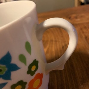 May include: White ceramic mug with a floral design featuring blue, green, orange, and yellow flowers. The mug has a white handle.