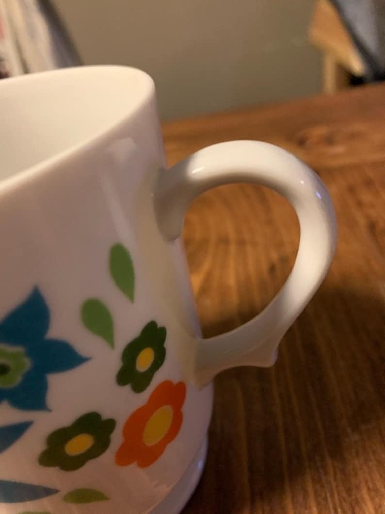 May include: White ceramic mug with a floral design featuring blue, green, orange, and yellow flowers. The mug has a white handle.
