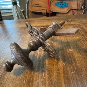 Pode incluir: Uma maçaneta de porta antiga de metal enferrujado com um botão redondo e placa traseira decorativa. A maçaneta é feita de metal envelhecido, mostrando sinais de desgaste. O design inclui uma peça retangular e uma haste roscada.