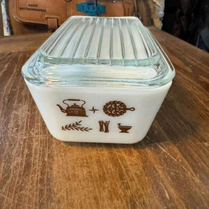 May include: A vintage white and clear glass casserole dish with a lid. The dish has a geometric shape and features brown kitchen-themed illustrations. The lid is clear with vertical ridges.