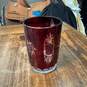 May include: A deep red glass tumbler featuring intricate cut-glass star and geometric patterns on its exterior. The cylindrical vessel has a clear, scalloped base and a smooth, open top rim, showcasing a vibrant ruby-colored translucent finish.