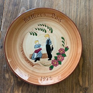 May include: A brown ceramic plate with a painted scene of two figures in a woodland setting. The plate has the words "Mother's Day 1972" written on it.