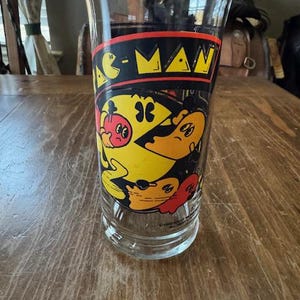 May include: A clear glass featuring a Pac-Man design. The design includes the words "PAC-MAN" in yellow and black, along with images of Pac-Man and ghosts. The glass is cylindrical and sits on a wooden surface.