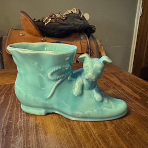 Puede incluir: Una maceta de cerámica turquesa con forma de bota con una figura de perro en el lateral. La bota tiene un detalle de lazo y un borde festoneado. La maceta está sobre una superficie de madera.