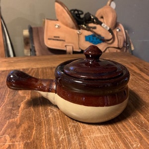 Puede incluir: Un olla de cerámica marrón y blanca con tapa y asa. La olla está sobre una superficie de madera.