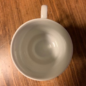 May include: A white ceramic teacup with a simple design. The cup is empty and has a handle on the right side.