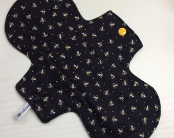 May include: Black reusable cloth pad with a bee print and a yellow snap button. The pad has a white label with the text "Miodera".