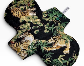 May include: A reusable cloth menstrual pad featuring a black background with a repeating pattern of tigers, green bamboo leaves, and pine-like foliage. The pad includes two green snap fasteners and a small white fabric tag labeled Rag-Kag.