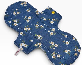 May include: A reusable cloth menstrual pad featuring a dark blue denim-look fabric with a white daisy floral pattern and small white polka dots. The pad has two white plastic snap buttons on one wing and a single yellow snap button on the other.