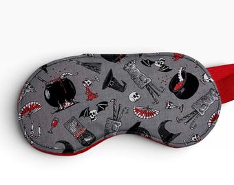 May include: A grey sleep mask featuring a repeating pattern of black cauldrons, witch hats, skulls, bats, red fans, and broomsticks. The mask has a red fabric trim and a matching red elastic strap attached to the side for securing it in place.