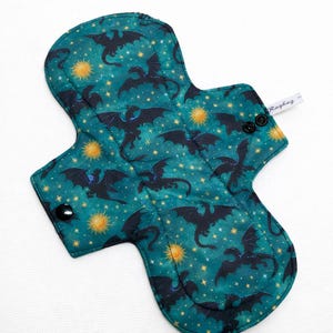 May include: A reusable cloth menstrual pad with a teal background featuring black dragon silhouettes, yellow suns, and white stars. The pad has a curved shape with snap closures and a small tag that says "Ragbag".
