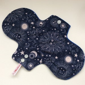 Puede incluir: Una compresa de tela reutilizable azul oscuro con un estampado celestial blanco con soles, lunas y estrellas. La compresa tiene un ribete azul y dos broches blancos. El texto "Madeira" está impreso en la compresa.