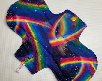 May include: A reusable cloth menstrual pad with a colorful rainbow and galaxy print. The pad has three snap closures and a label that says "Heas".