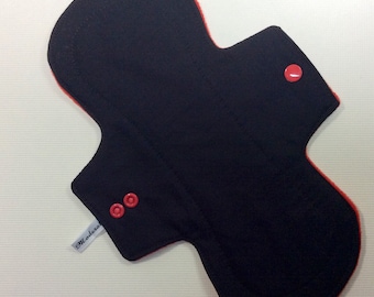 May include: Black reusable cloth menstrual pad with red trim and red snap buttons. The pad has a white label that says "Moderna".