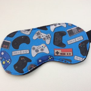 Könnte beinhalten: Eine blaue Schlafmaske mit einem Muster aus verschiedenen Videospiel-Controllern in Schwarz, Grau und Rot. Die Maske hat ein schwarzes Gummiband und ein weiches, schwarzes Futter. Sie ist zum Abdunkeln konzipiert.