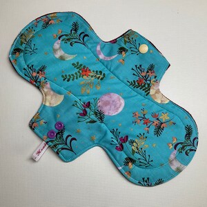 May include: A turquoise blue reusable cloth menstrual pad with a floral and moon print. The pad has two purple snaps and a white tag that says "Heaux".