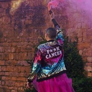 May include: A person wearing a sequined jacket with the text "F**K CANCER" on the back, paired with a pink tulle skirt. The jacket has a rainbow colour scheme. The person is holding a pink smoke flare.