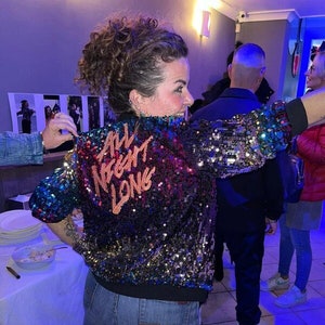May include: A woman wearing a black sequined jacket with the text "All Night Long" in pink, blue, and orange. She is also wearing a denim skirt and black boots.