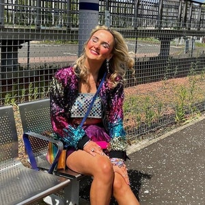 May include: A colorful sequined jacket with a rainbow of colors, paired with a checkered crop top and a pink skirt. The outfit is completed with pink sneakers with brown soles. A blue purse hangs from the shoulder.