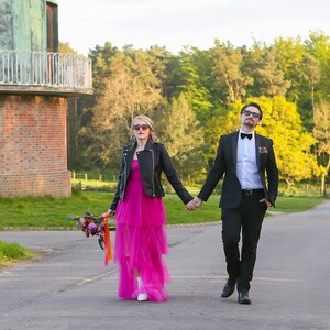 May include: A couple walking on a paved road, holding hands. The woman wears a black leather jacket over a bright pink tiered dress and white boots. The man wears a black suit with a bow tie. The woman carries a bouquet of flowers.