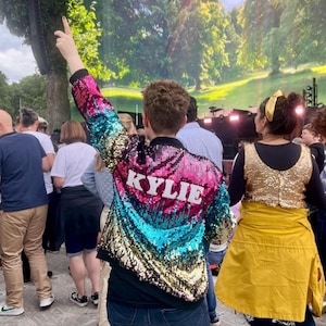 May include: A person wearing a multi-colored sequined jacket with the word "KYLIE" written on the back. The jacket has pink, blue, and gold sequins. The person is standing in a crowd of people.