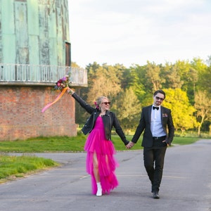 May include: A couple walks down a road, holding hands. The woman wears a hot pink dress, black leather jacket, and white boots. The man wears a black suit with a bow tie. The woman holds a bouquet of flowers.