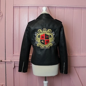 Pu&ograve; includere: Giacca da motociclista nera in ecopelle con stemma ricamato sul retro. Lo stemma presenta un'impronta di zampa, una fetta di pizza e un design a pentagramma. Include dettagli con cerniera e colletto a revers.