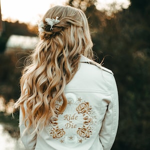 May include: Cream-colored jacket with intricate embroidery on the back, featuring roses, moons, stars, and the words "Ride or Die." The jacket is worn by a person with long, wavy blonde hair, styled with a floral hair accessory.