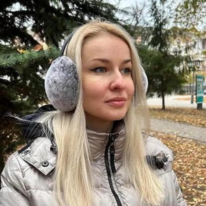 May include: A woman wearing a gray puffer jacket with a black zipper and a pair of gray faux fur earmuffs.