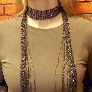 Puede incluir: Un collar choker de lentejuelas marrones con largos mechones colgantes. El choker es una banda ancha, y los mechones caen en cascada por el frente. El collar se usa sobre una camisa color canela, creando un contraste de texturas y colores.