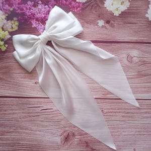 May include: A large white satin bow with long tails. The bow is tied in a knot and the tails are flowing down. The bow is on a wooden surface.
